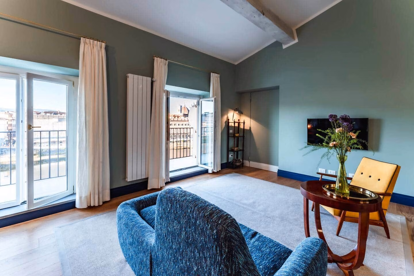 Luxury View Arno — soggiorno verde salvia con divano chenille blu e porte-finestre sul balcone