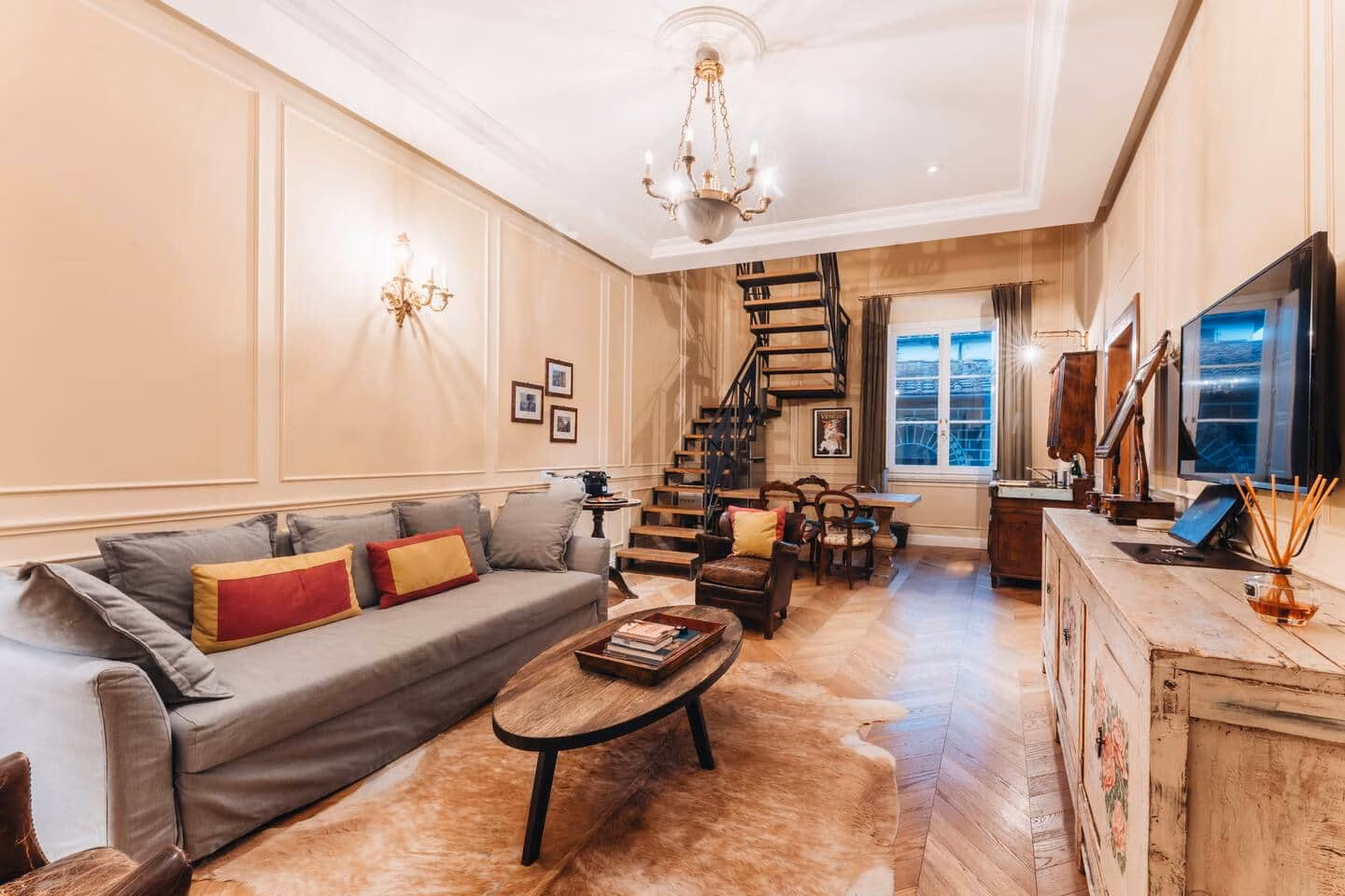 Le Ruote — soggiorno con boiserie, divano in lino grigio, lampadario e scala nera a vista su parquet a spina di pesce